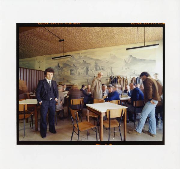Interno. Bar - gruppo di uomini - uomo in piedi - tavolini - sedie - quadro di paesaggio