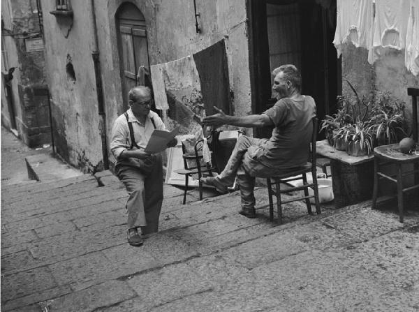 Napoli: Incontri scontri. Napoli - Strada, scalinata - Ritratto maschile - Anziano seduto su una sedia sull'uscio di casa e anziano con fogli di carta - Gesti, lettura