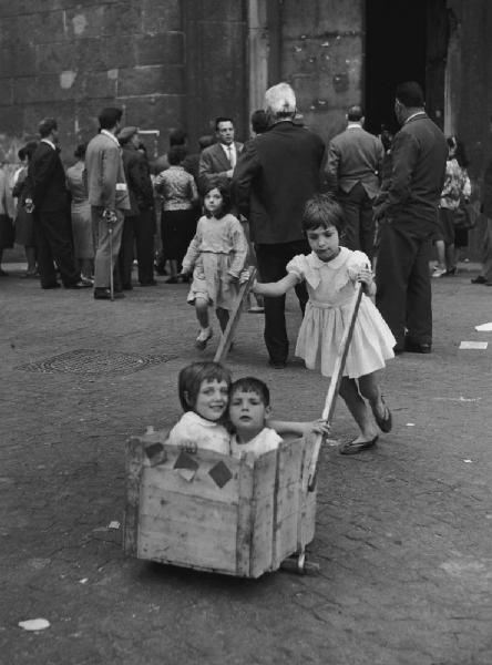 Napoli: Scene di vita varie. Napoli - Strada - Ritratto di gruppo - Due bambini dentro una carriola artigianale di legno e una bambina spinge - Gioco - Folla sullo sfondo