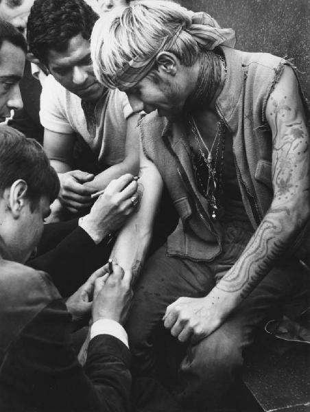 Mondo Beat. Milano - Piazza del Duomo - Monumento a Vittorio Emanuele II - Ritratto di gruppo - Ragazzi disegnano tatuaggi sulle braccia di un ragazzo con bandana e gilet