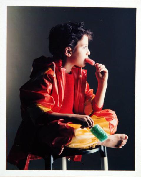 Campagna pubblicitaria per Trussardi Junior - Bambino seduto di profilo: mantella e pantaloni cerati rossi e gialli - Due ghiaccioli in mano