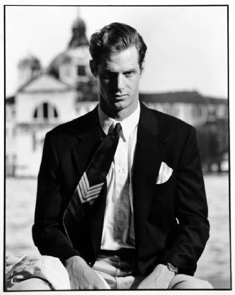 Campagna pubblicitaria per Trussardi Uomo - Modello seduto: giacca scura su pantaloni a righe, camicia bianca e cravatta regimental