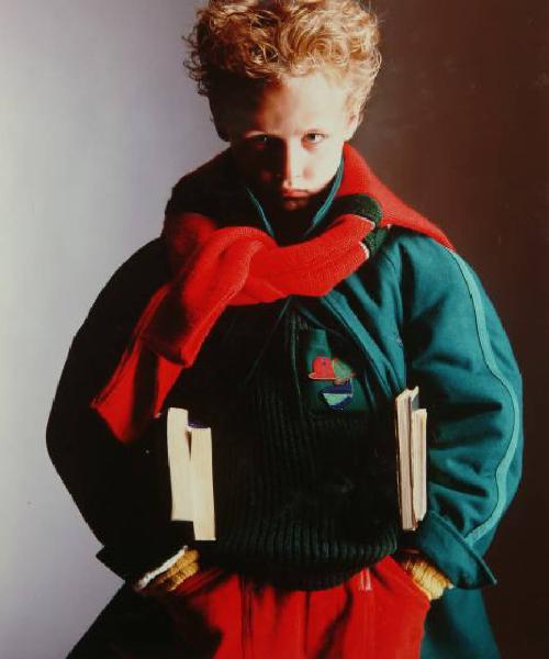 Campagna pubblicitaria per Trussardi Junior - Bambino con mani in tasca: cappotto di panno verde, maglione a coste, pantaloni di velluto rossi e cardigan rosso annodato attorno al collo - Libri