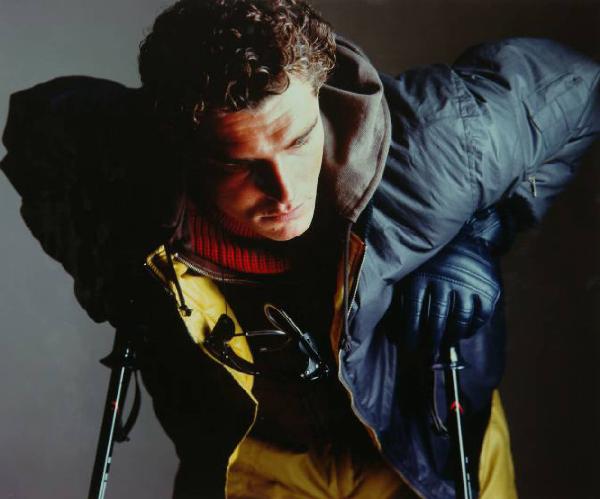 Campagna pubblicitaria per Trussardi Uomo - Sportswear - Modello appoggiato a racchette da sci: giacca a vento blu su pantaloni gialli, guanti di pelle blu - Sciatore