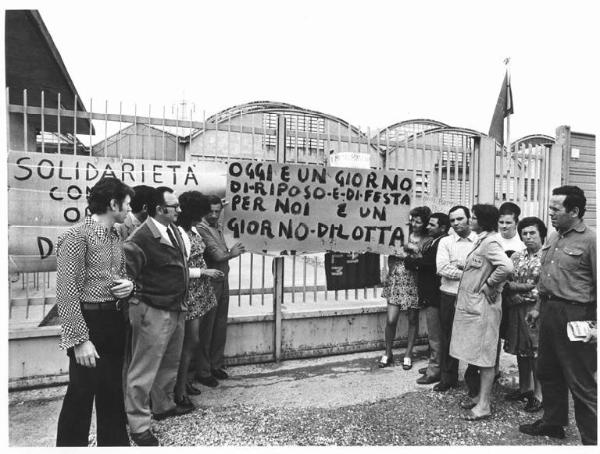 Ago Articoli Gomma - Fabbrica occupata - Lavoratori davanti ai cancelli della fabbrica - Operaia con grembiule da lavoro - Striscione - Bandiera