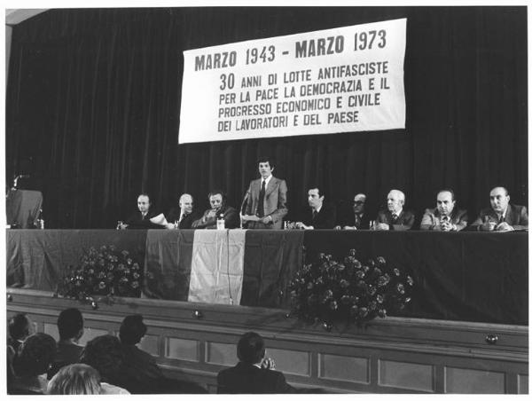 Teatro Odeon - Interno - Commemorazione degli scioperi del marzo 1943 - Antifascismo - Tavolo della presidenza con tra gli altri G. Polotti, R. Romei, L. De Carlini, P. Perotta, A. Bonaccini - M. Colombo parla alla platea - Parola d'ordine - Bandiera