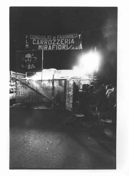 Fabbrica Fiat - Picchetto notturno dei lavoratori alle porte 3-4-5 contro i licenziamenti - Lavoratori davanti all'ingresso - Striscione Flm - Bandiera con volto di Karl Marx - Scritte di lotta