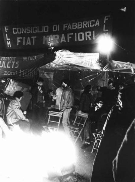Fabbrica Fiat - Picchetto notturno dei lavoratori alle porte 3-4-5 contro i licenziamenti - Lavoratori davanti all'ingresso intorno al fuoco - Striscione Flm