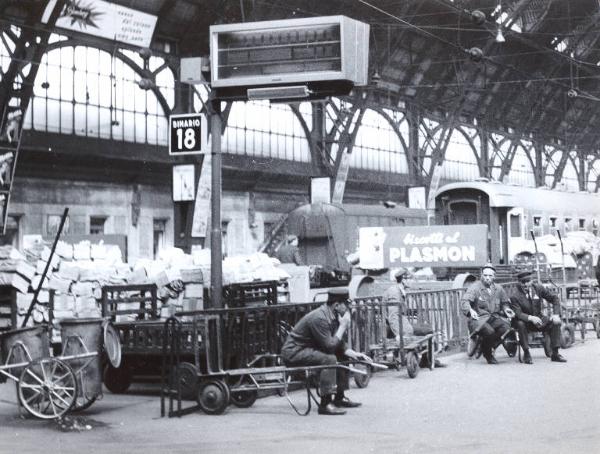 Milano - Sciopero ferrovieri - Stazione Centrale - Interno - Binari - Facchini in divisa seduti sui carrelli - Carrelli pieni di posta - Cartello pubblicitario dei biscotti Plasmon