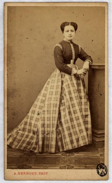 Ritratto femminile - Giovane donna con gonna a scacchi, in piedi appoggiata a un piedistallo.