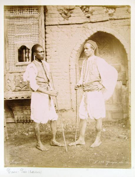 Ritratto di gruppo - Due uomini a piedi scalzi con in mano un bastone posano davanti alla facciata di una casa del Cairo con il tipico balcone ligneo aggettante chiamato moucharabia