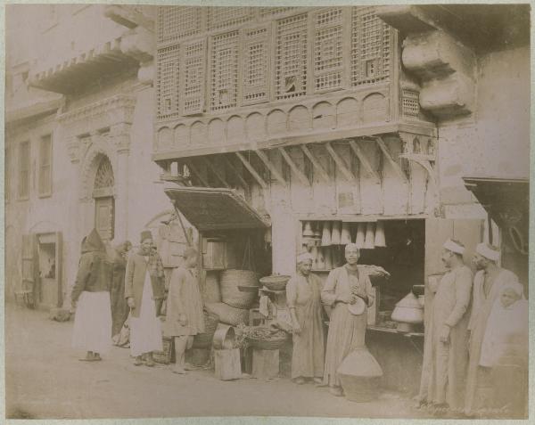 Egitto - Il Cairo - Strada - Bazar - Bottega - Drogheria - esposizione della merce sulla strada - Due uomini guardano in camera - Passanti - Balcone ligneo aggettante chiamato moucharabia