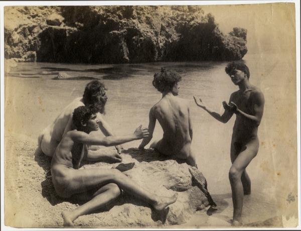 Quattro nudi maschili, tre seduti su uno scoglio e uno in piedi nel mare