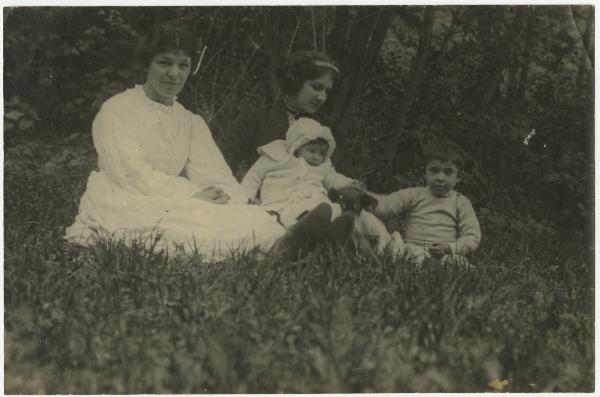 Ritratto di famiglia - Elvira Lazzaroni seduta con la figlia Leopolda Metlicovitz sulle gambe, il figlio Roberto Metlicovitz e altra donna - Esterno, prato - Alberi sullo sfondo