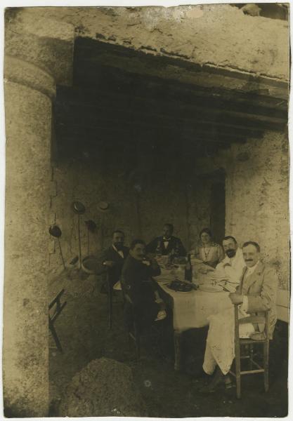 Ritratto di gruppo - Elvira Lazzaroni e Leopoldo Metlicovitz con altri uomini seduti a una tavola imbandita - Pasto - Esterno, porticato rustico