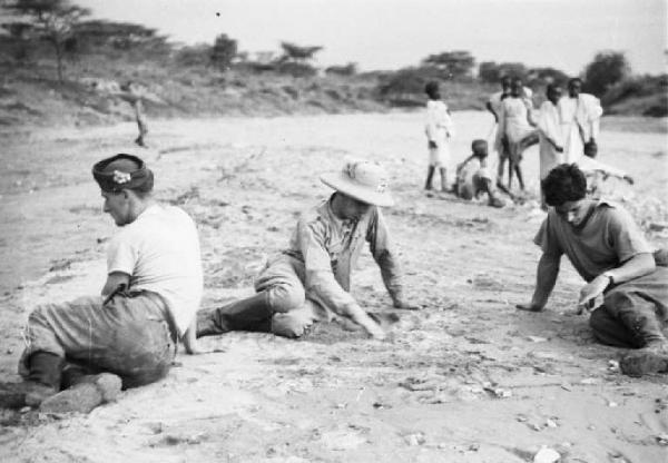 Viaggio in Africa. Tre militari itliani si riposano seduti a terra. Sullo sfondo un gruppo di indigeni