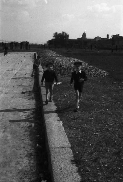 Periferia di Milano: bambini corrono lungo il ciglio della strada. In lontananza i caseggiati
