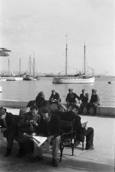 Viaggio in Jugoslavia. Sebenico: gruppo di abitanti chiaccherano seduti sulle panchine al porto - sullo sfondo il mare