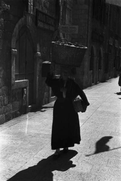Viaggio in Jugoslavia. Dubrovnik (Ragusa): scene di vita quotidiana - donna che cammina lungo una strada della cittadina portando sulla testa una cesta