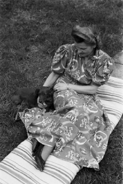 Internamento in Svizzera. Attisholz. Ritratto femminile, Milly posa nel giardino di casa mentre accarezza un cane bassotto. Indossa un vistoso abito fantasia