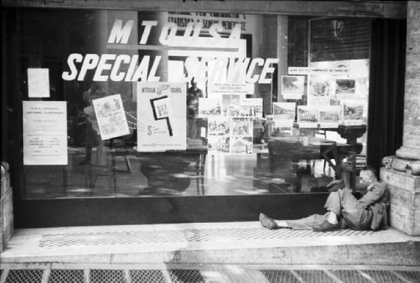 Italia Dopoguerra. Roma - Uomo sdraiato davanti alla vetrina di un esercizio pubblico recante la scritta "mtousa special service"