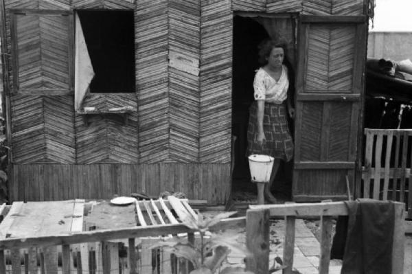 Italia Dopoguerra. Milano - Periferia - Baraccopoli - Donna che esce da una baracca trasportando un secchio d'acqua