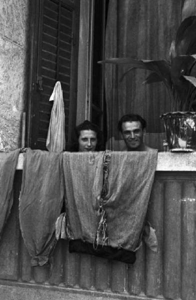 Italia Dopoguerra. Periferia di Milano, le case popolari del quartiere "Baia del Re". Ritratto di coppia, un uomo e una donna che osservano da un balcone sul cui parapetto sono stesi dei pantaloni ad asciugare