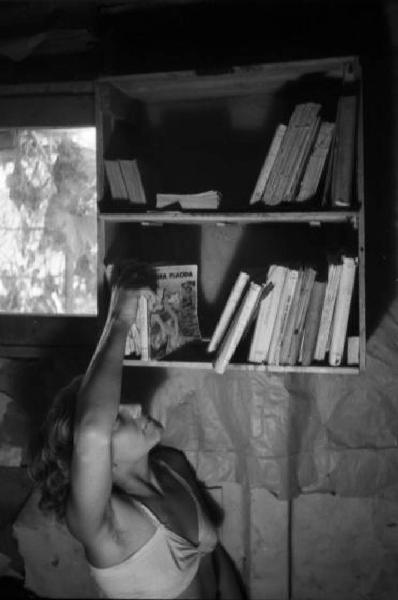 Italia Dopoguerra. Milano - Periferia - Baraccopoli - Norma Barbieri sceglie un libro dalla propria biblioteca di fortuna