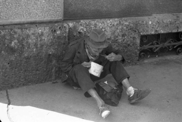 Italia Dopoguerra. Milano - Un uomo seduto su un marciapiede consuma il proprio pasto mangiando da una scodella