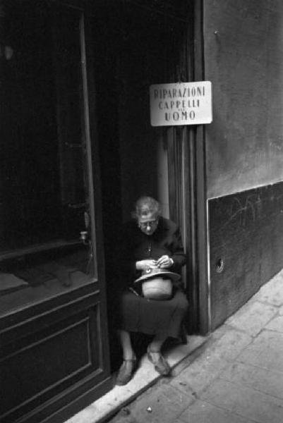 Italia Dopoguerra. Genova - Donna ripara un cappello seduta sulla soglia del proprio negozio