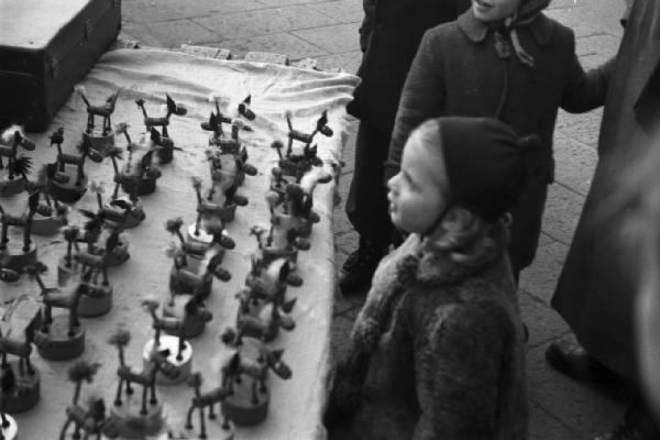 Italia Dopoguerra. Milano - fiera degli Obèi - Obèi: una bimba davanti a una bancarella di piccoli pupazzi mentre si rivolge al venditore