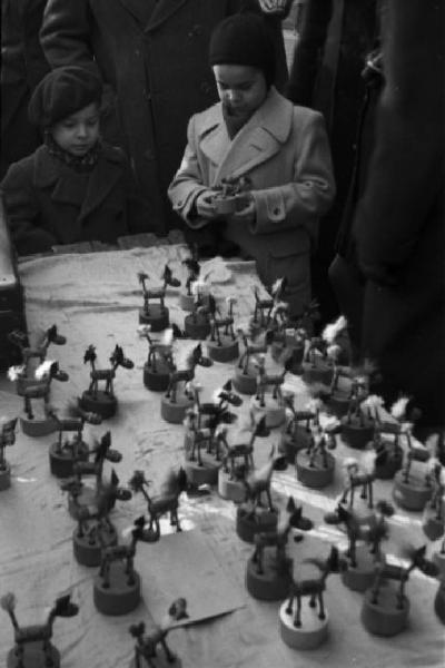 Italia Dopoguerra. Milano - fiera degli Obèi - Obèi: una coppia di bambini mentre prende una serie di giocattoli sopra una bancarella