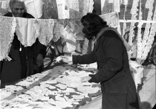 Italia Dopoguerra. Milano - fiera degli Obèi - Obèi: una donna sorpresa di spalle durante l'acquisto di merce presso un venditore di centrini lavorati a mano