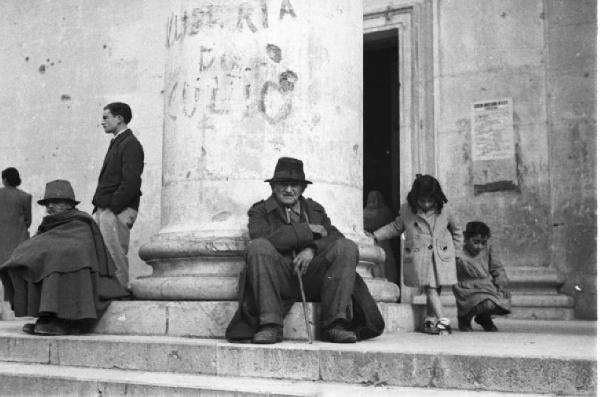 Italia Dopoguerra. Terracina. Due anziani seduti sulle gradinate di un edificio in prossimità di una colonna e due bambine
