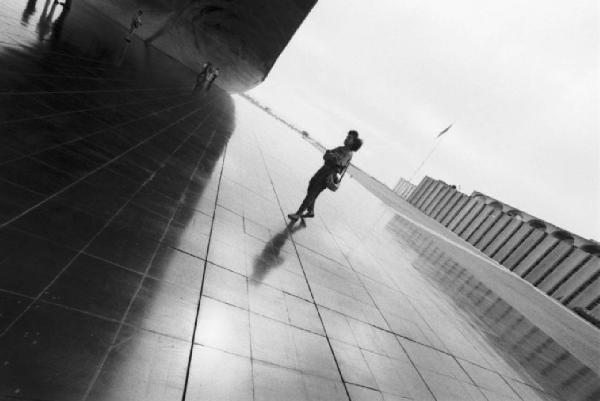 Brasilia. Piazzale lastricato con struttura curva in calcesruzzo e palazzo sullo sfondo. Si nota una figura femminile (Cyssa)