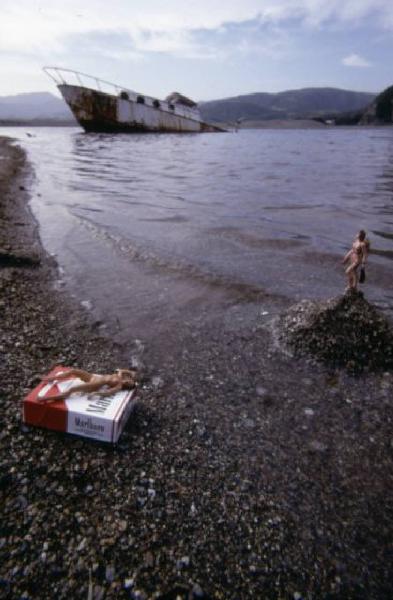 Marlboro. Pacchetto di sigarette sul bagnasciuga, con piccole bambole nude intorno e relitto di nave sullo sfondo