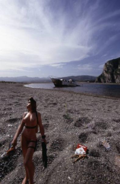 Marlboro. Pacchetto di sigarette sulla spiaggia, con piccole bambole nude intorno e relitto di nave sullo sfondo