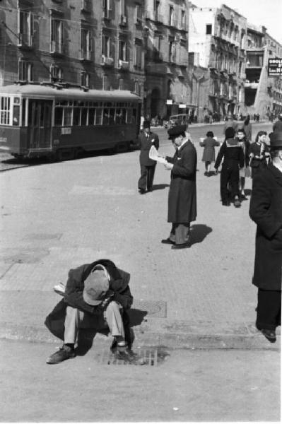 Italia Dopoguerra. Napoli. Passanti per le vie della città. Un uomo siede sul bordo del marciapiede. Un tram passa sullo sfondo