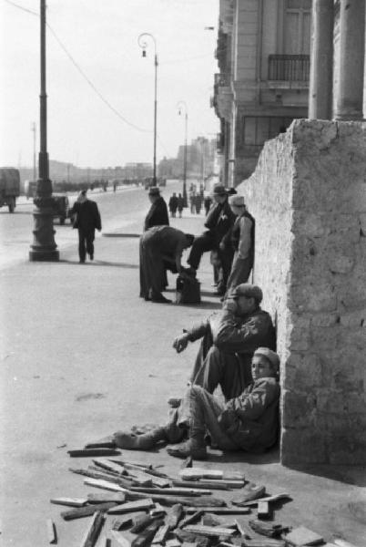 Italia Dopoguerra. Napoli. Veduta di via Caracciolo: due persone sedute per terra e un lustrascarpe. Automezzi lungo la strada