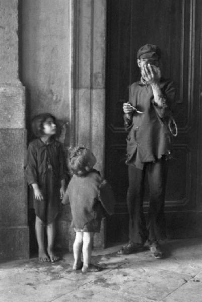 Italia Dopoguerra. Palermo. Nei pressi della stazione ferroviaria. Due bambine scalze con il padre dagli abiti stracci mentre mangia da un barattolo