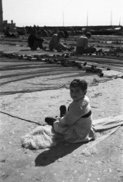 Italia Dopoguerra. Mondello. Italia Dopoguerra. Mondello. Ritratto di una bambina seduta per terra mentre i pescatori sistemano le reti