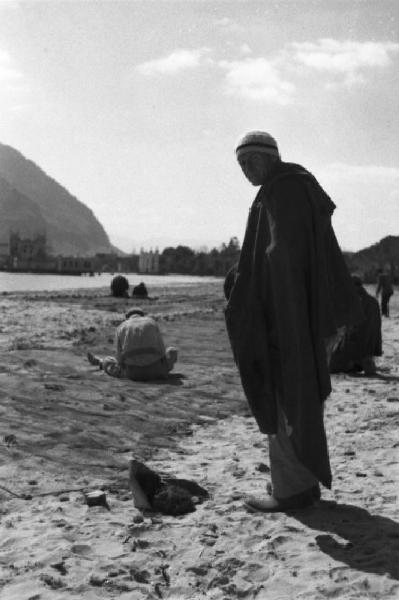 Italia Dopoguerra. Mondello. Ritratto maschile, un uomo in piedi sulla spiaggia mentre dei pescatori sistemano le reti