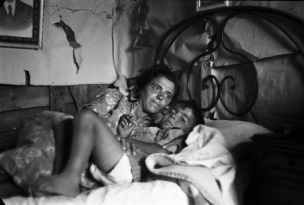 Italia Dopoguerra. Milano - Periferia - Baraccopoli - Ritratto di coppia, madre e figlio stesi sul letto all'interno di una baracca