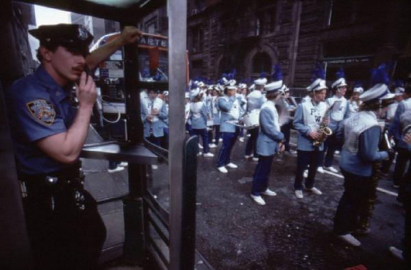 New York. Manhattan - banda sfila per le strade cittadine. Poliziotto parla da un telefono pubblico