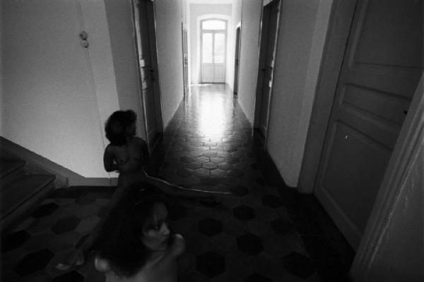 Due figure femminili lungo il corridoio di un'abitazione, una seduta a terra, l'altra ritratta in primo piano