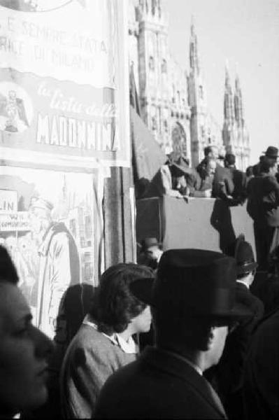 Milano. Piazza Duomo. Gente assiepata attorno e sopra il palco comizi - in primo piano un manifesto di propaganda elettorale - sullo sfondo il Duomo