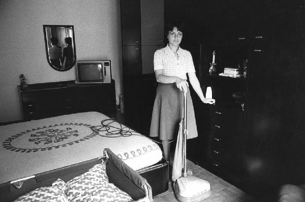 Una, nessuna, centomila. La Casa e i Riti. Milano - Camera da letto, interno - Televisore - Ritratto femminile: donna con aspirapolvere