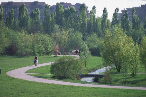 Sesto San Giovanni - Parco Nord, settore Montagnetta - Percorso ciclopedonale che costeggia il canale del laghetto artificiale Suzzani - Ponticello - Persone che corrono e a passeggio - Bambino e adulto in bicicletta - Filari di alberi - Case