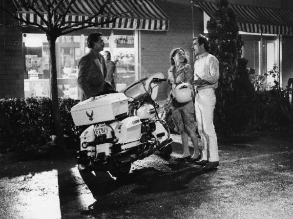 Scena del film "La cicala" - Regia Alberto Lattuada - 1980 - Gli attori Anthony Franciosa, Barbara De Rossi e Antonio Catanfora accanto a una moto. Alle loro spalle Virna Lisi