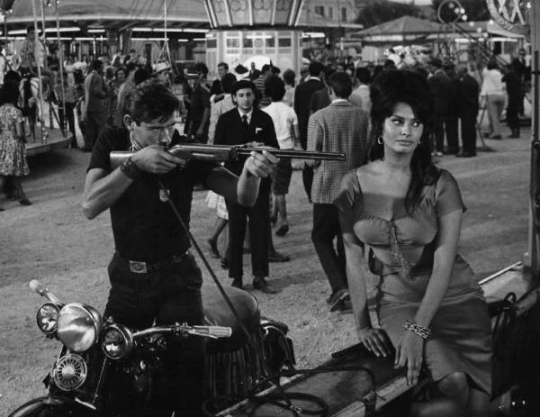 Scena dell'episodio "La riffa" del film "Boccaccio '70" - Regia Vittorio De Sica - 1962 - L'attrice Sophia Loren e un attore non identificato che con fucile gioca a tiro al bersaglio alle giostre
